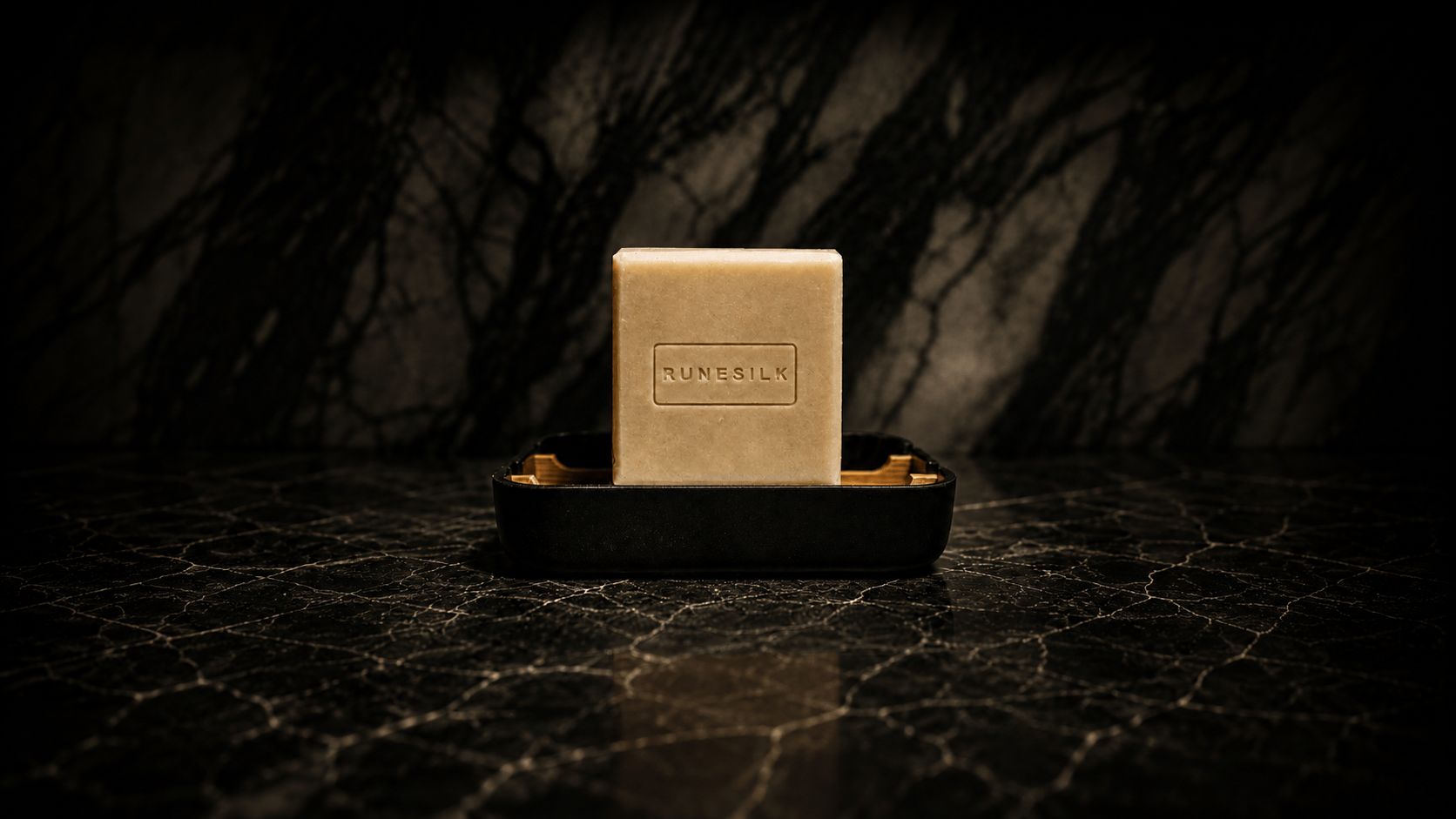 A square beige bar of soap or shampoo block standing upright in a black rectangular holder on a dark marble-like surface, with a shadowy tree silhouette in the background. The front of the block shows the embossed brand name RUNESILK.